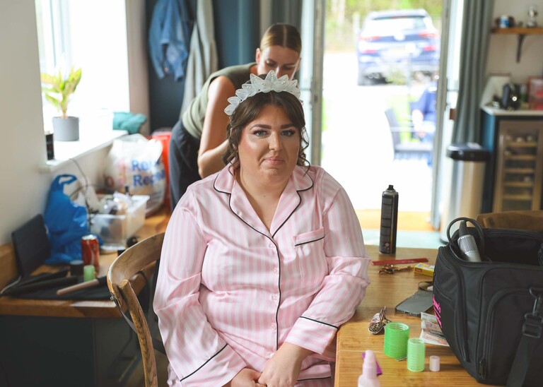 A bride sat in silk light pink pjs having her hair done and head dress put on on a wedding day at Hungarian Hall Estate captured by Suffolk Wedding Photographer Hayley Denston Photography