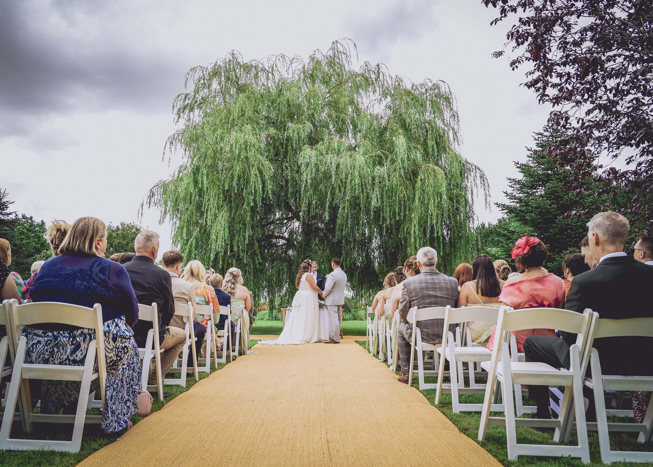 A bride and groom holding hands from behind with a big willow tree taken during their outdoor wedding ceremony on a wedding day at Hungarian Hall Estate captured by Suffolk Wedding Photographer Hayley Denston Photography