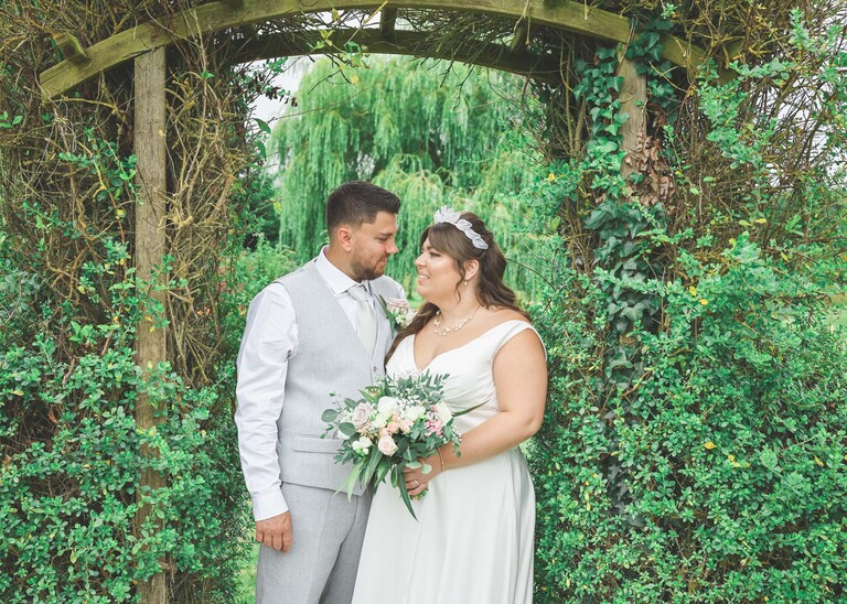 A bride and groom under a wooden archway covered with climbers looking at each other on a wedding day at Hungarian Hall Estate captured by Suffolk Wedding Photographer Hayley Denston Photography
