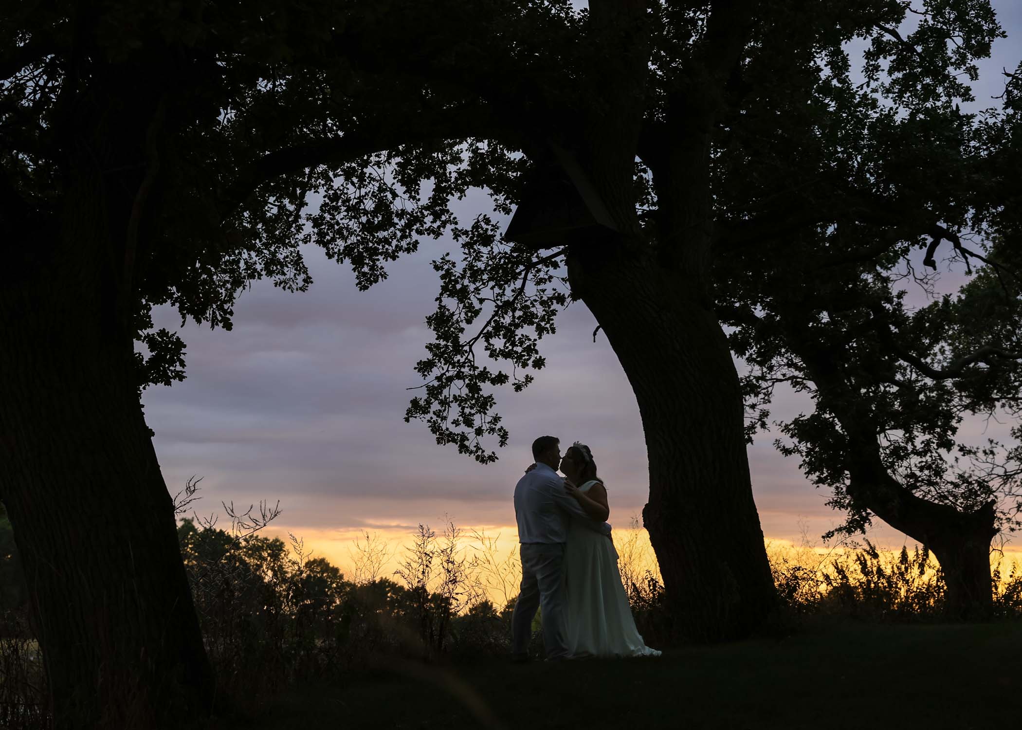 A bride and groom silloueted with trees in an archway and sunset behind them on a wedding day at Hungarian Hall Estate captured by Suffolk Wedding Photographer Hayley Denston Photography
