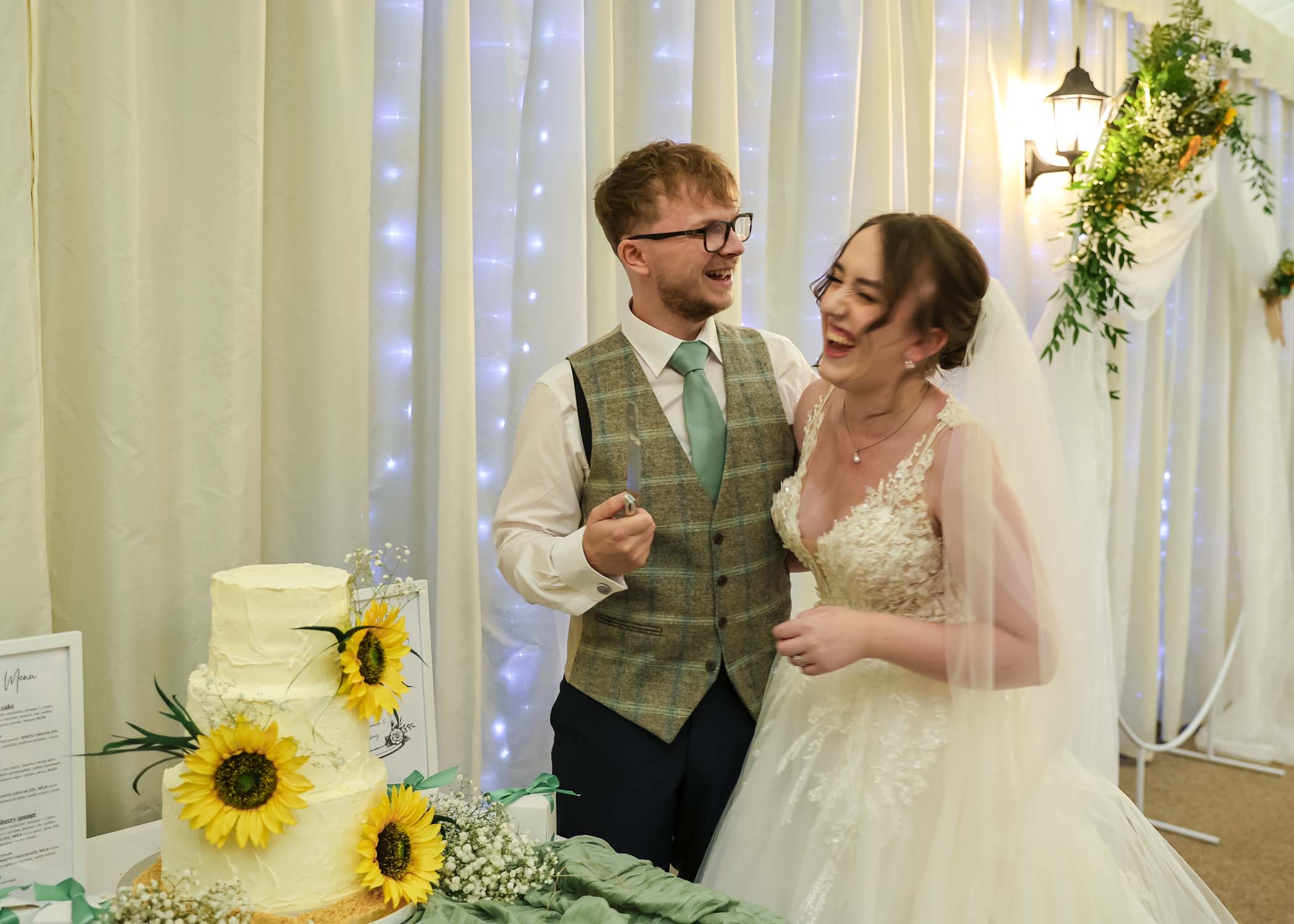 A bride and groom laughing as they cut their sunflower wedding cake on a wedding day at Applewood Hall in Norfolk captured by suffolk and norfolk wedding photographer Hayley Denston Photography