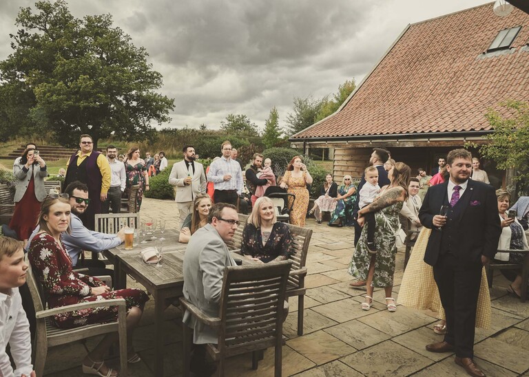 Guests sat outdoors in a courtyard area listening to wedding speeches on a wedding day at Easton Grange captured by Suffolk Wedding Photographer Hayley Denston Photography