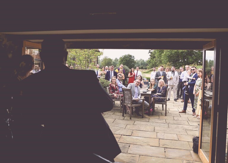 A silouetted father of the bride with guests outside watching during wedding speeches on a wedding day at Easton Grange captured by Suffolk Wedding Photographer Hayley Denston Photography