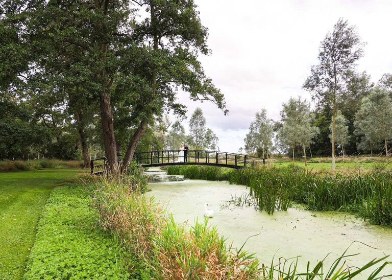 A long shot of a bride and groom stood on a bridge surrounded by trees and the river on a wedding day at Easton Grange captured by Suffolk Wedding Photographer Hayley Denston Photography 