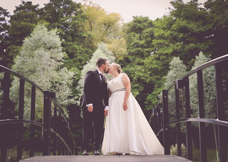 A bride and groom stood on a bridge kissing looking up the bridge on a wedding day at Easton Grange captured by Suffolk Wedding Photographer Hayley Denston Photography 