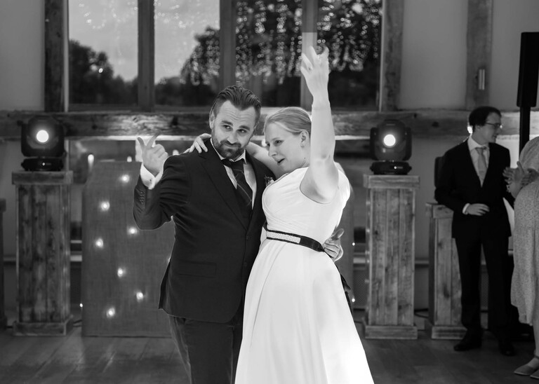 A black and white photo of a bride and groom at the end of their first dance with their arms in the air encouraging guests onto the dance floor on a wedding day at Easton Grange captured by Suffolk Wedding Photographer Hayley Denston Photography 