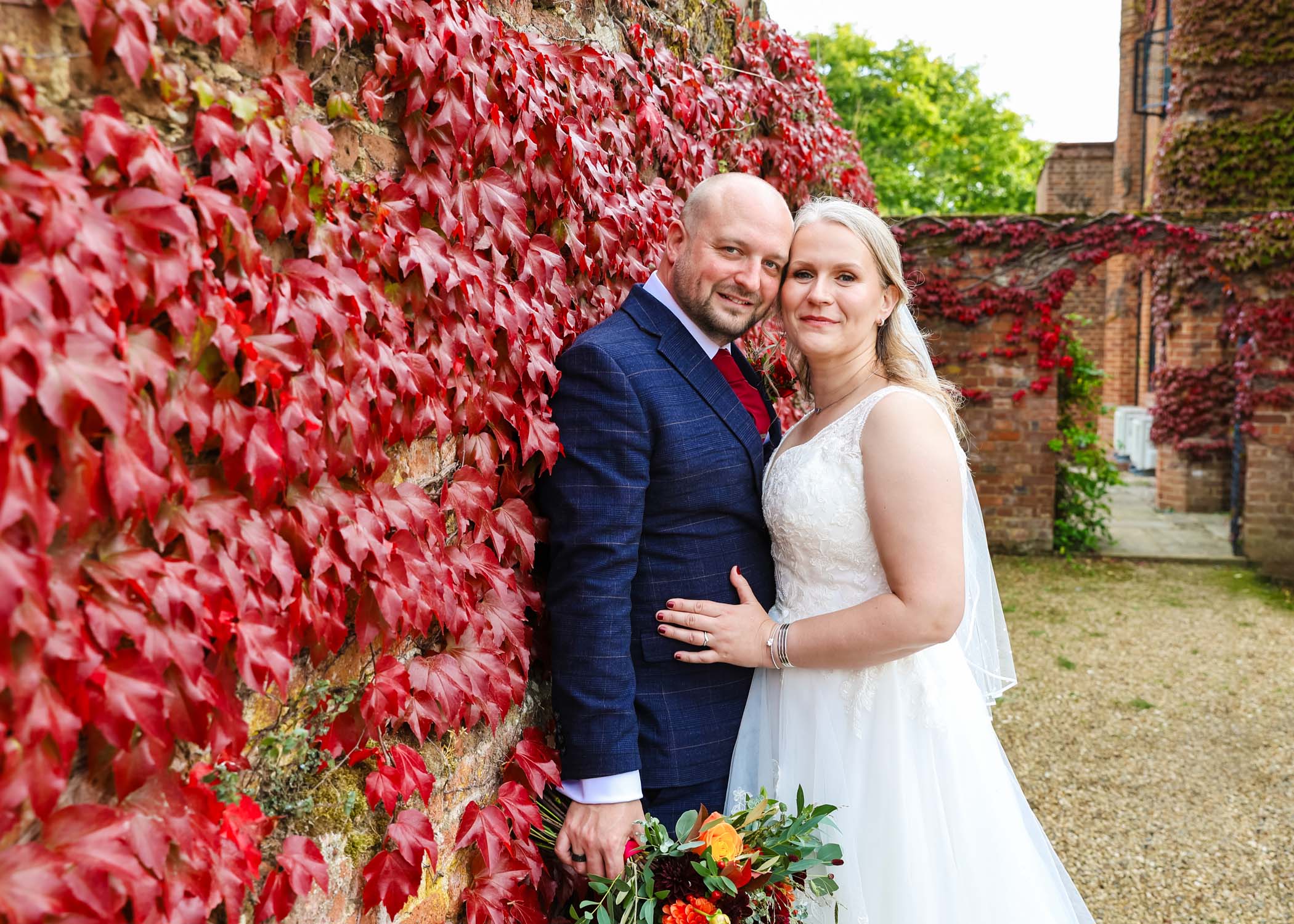 A bride and groom stood hugging each other along a wall of bright red leaf climbers on a wedding day at Seckford Hall photographed by Suffolk Wedding Photographer Hayley Denston Photography