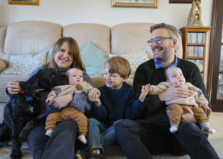 A family of two adults, a small boy, twins and a dog licking one of the babies sat on the floor in front of a sofa captured by Suffolk Portrait Photographer Hayley Denston Photography