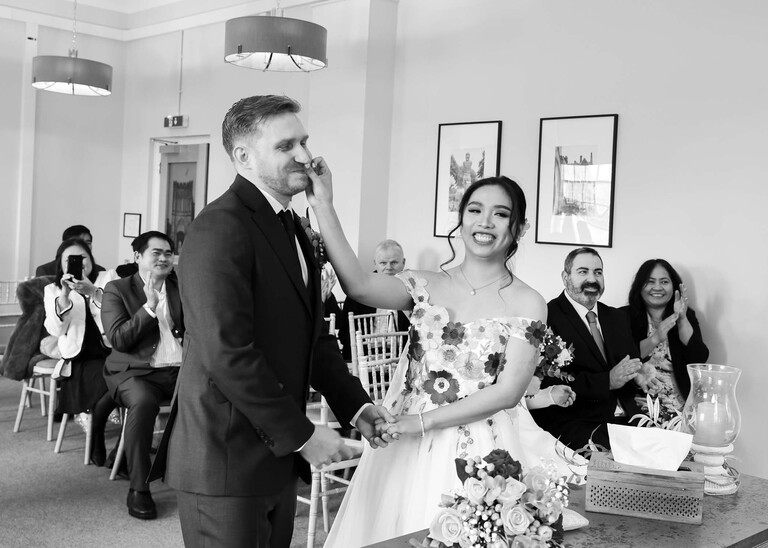 A black and white photo of a bride wiping lipstick off of the grooms cheek during their wedding ceremony at Bury St Edmunds Registry Office photographed by Suffolk Wedding Photographer Hayley Denston Photography