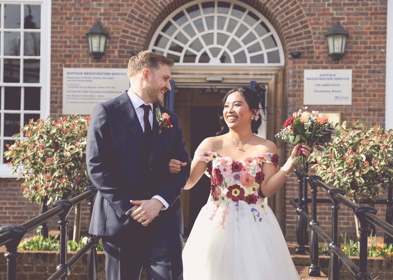 A bride and groom walking out of the registry office smiling and looking happy. The bride is wearing a bright floral embroidered wedding dress at Bury St Edmunds Registry Office photographed by Suffolk Wedding Photographer Hayley Denston Photography