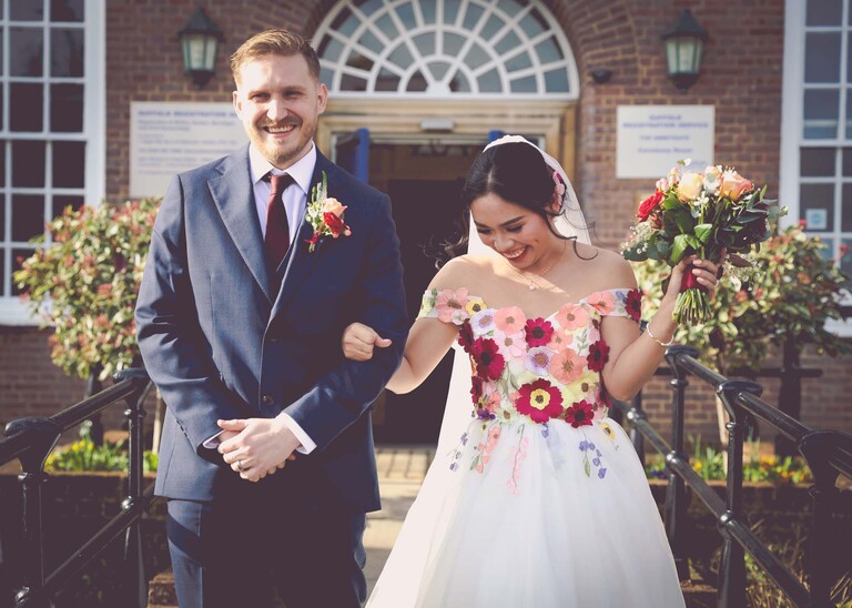 A bride and groom walking out of the registry office smiling and looking happy. The bride is wearing a bright floral embroidered wedding dress at Bury St Edmunds Registry Office photographed by Suffolk Wedding Photographer Hayley Denston Photography