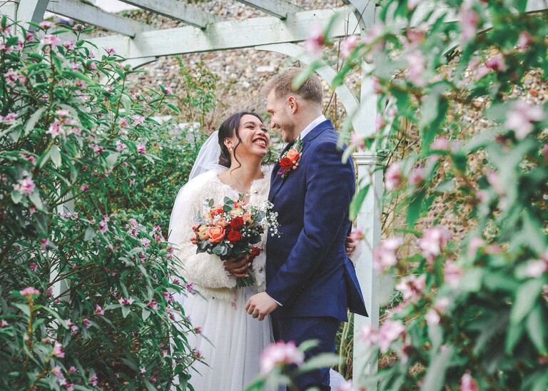 A bride and groom stood under a green arbour with daphne flower bushes in bloom on their winter wedding day in Abbey Gardens in Bury St Edmunds on a wedding day photographed by Suffolk Wedding Photographer Hayley Denston Photography