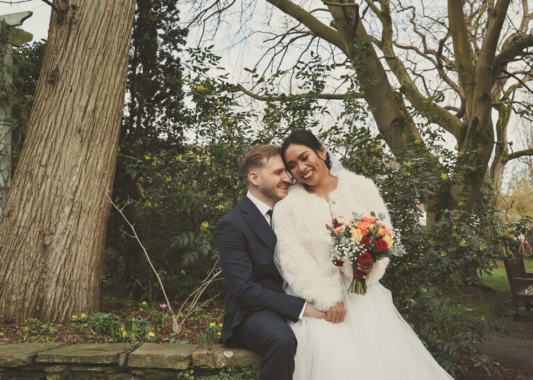 A bride sat on her grooms lap snuggling in to each other smiling on their winter wedding day in Abbey Gardens in Bury St Edmunds on a wedding day photographed by Suffolk Wedding Photographer Hayley Denston Photography