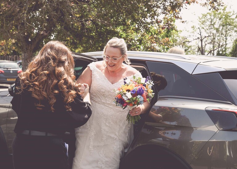 A bride with her bright rainbow coloured bouquet getting out of a car on a wedding day at Venue 16 captured by suffolk wedding photographer Hayley Denston Photography