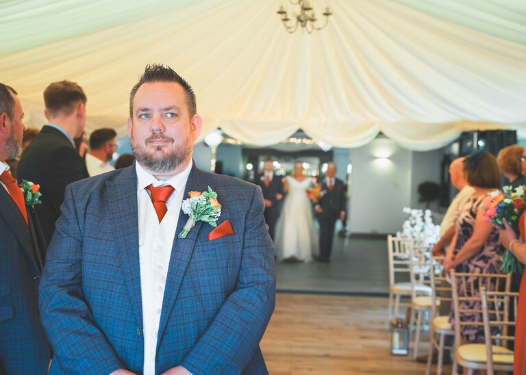A groom looking apprehensively at the registrar for the nod to turn round for a wedding ceremony as his bride walks in on a wedding day at Venue 16 captured by suffolk wedding photographer Hayley Denston Photography