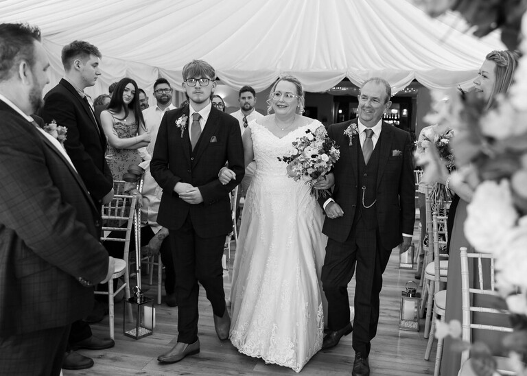 A black and white photo of a bride being walked down the aisle by her son and dad on a wedding day at Venue 16 captured by suffolk wedding photographer Hayley Denston Photography