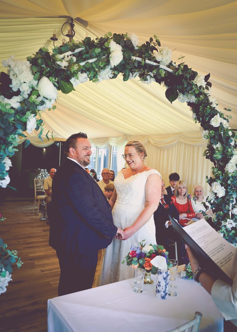 A bride and groom through a flower arch with white roses during their wedding ceremony on a wedding day at Venue 16 captured by suffolk wedding photographer Hayley Denston Photography