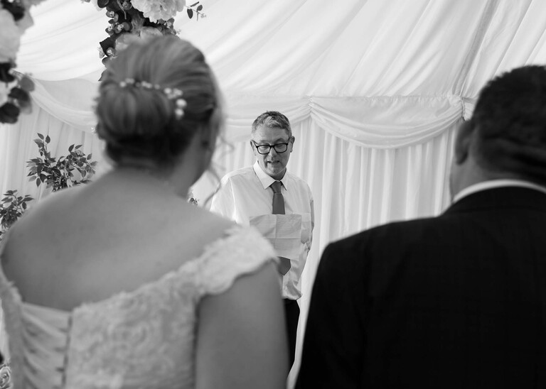 A black and white photo taken over the shoulder of a bride and groom at a speaker giving a reading during their wedding ceremony on a wedding day at Venue 16 captured by suffolk wedding photographer Hayley Denston Photography