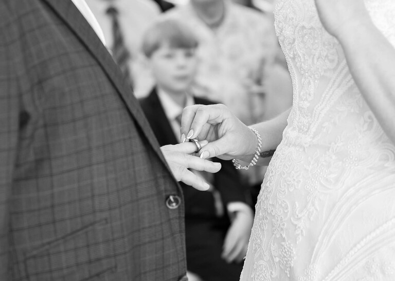 A black and white photo of a bride placing the ring on the grooms finger during their wedding ceremony on a wedding day at Venue 16 captured by suffolk wedding photographer Hayley Denston Photography