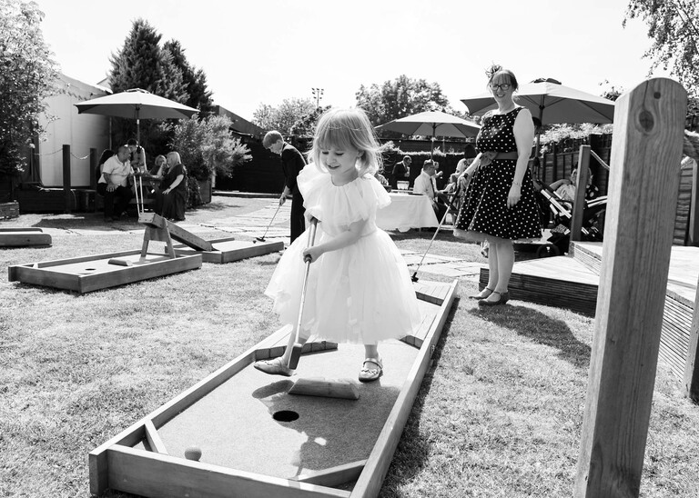 A black and white photo of a little flowergirl playing crazy golf at a wedding reception on a wedding day at Venue 16 captured by suffolk wedding photographer Hayley Denston Photography
