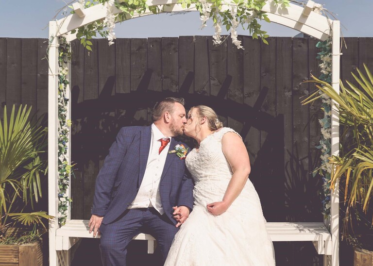 A bride and groom kissing sat on a seat with black fencing behind them and white wisteria on an archway around them on a wedding day at Venue 16 captured by suffolk wedding photographer Hayley Denston Photography