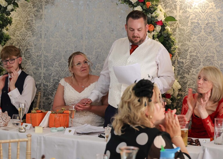 A groom getting emotional during his wedding speech and the bride holding his hand and looking up at him on a wedding day at Venue 16 captured by suffolk wedding photographer Hayley Denston Photography