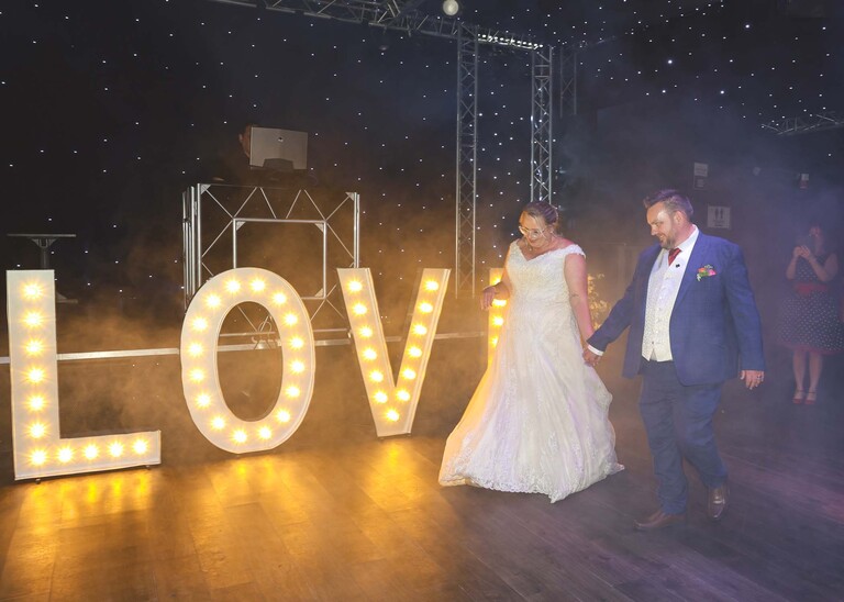 A bride and groom walking past giant lit love letters onto the dance floor for their first dance on a wedding day at Venue 16 captured by suffolk wedding photographer Hayley Denston Photography