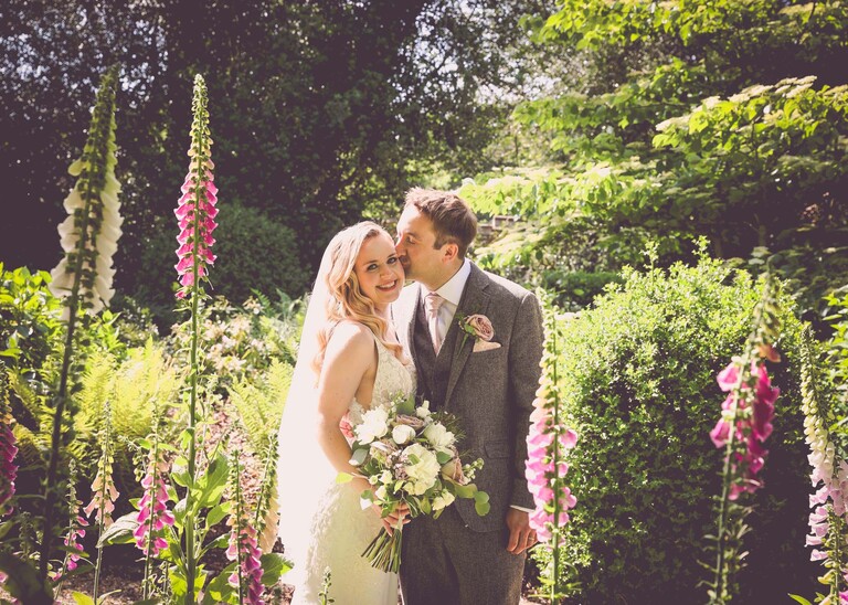 A groom kissing a bride on her head in amongst pink foxgloves with the sun behind them on a wedding day at Haughley Park Barn photographed by suffolk wedding photographer Hayley Denston Photography