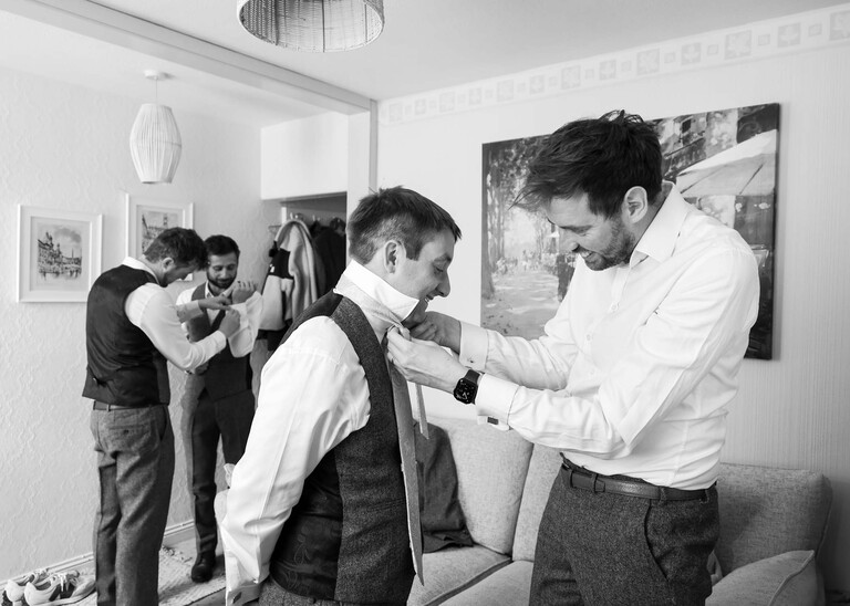 A black and white photo of men including the groom getting ready and helping each other put on ties on a wedding day at Haughley Park Barn photographed by suffolk wedding photographer Hayley Denston Photography
