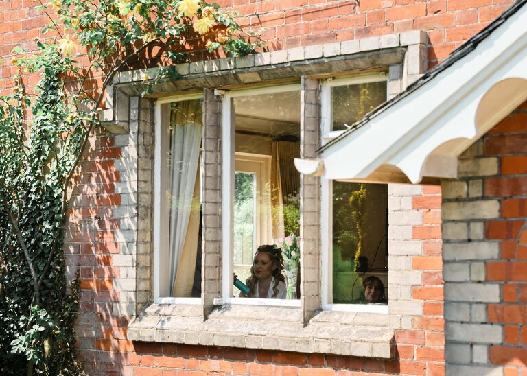 A bride through a farmhouse window having her hair and make up done on a wedding day at Haughley Park Barn photographed by suffolk wedding photographer Hayley Denston Photography