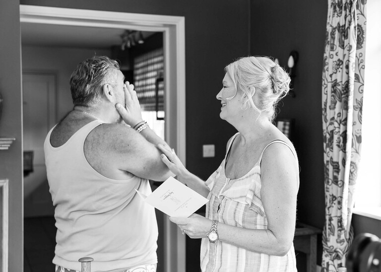 A black and white photo of a father of the bride crying and being comforted by his wife after a bride gives him a card and present on a wedding day at Haughley Park Barn photographed by suffolk wedding photographer Hayley Denston Photography