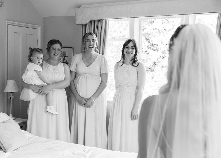 A black and white photo of bridesmaids looking at the bride for a first reveal of her wedding dress on a wedding day at Haughley Park Barn photographed by suffolk wedding photographer Hayley Denston Photography