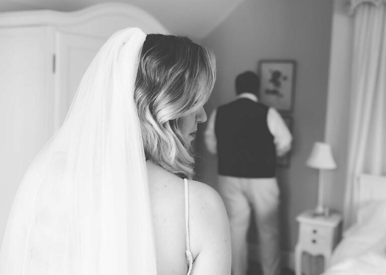A bride glancing to the sad in black and white with her dad in the background on a wedding day at Haughley Park Barn photographed by suffolk wedding photographer Hayley Denston Photography