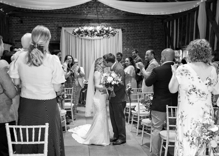 A black and white photo of a bride and groom kissing half way down the aisle as they make their exit on a wedding day at Haughley Park Barn photographed by suffolk wedding photographer Hayley Denston Photography