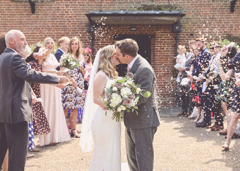 A bride and groom kissing in a confetti aisle as they are showered with confetti on a wedding day at Haughley Park Barn photographed by suffolk wedding photographer Hayley Denston Photography