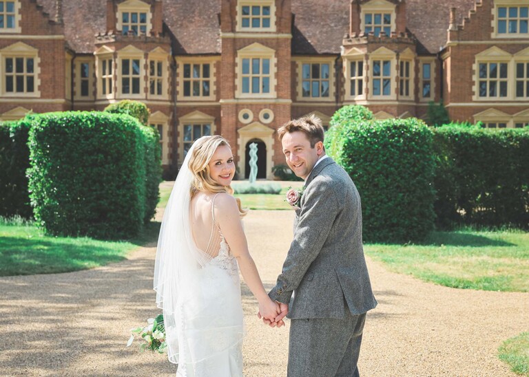 A bride and groom holding hands and looking back over their shoulders with a manificent hall behind them on a wedding day at Haughley Park Barn photographed by suffolk wedding photographer Hayley Denston Photography