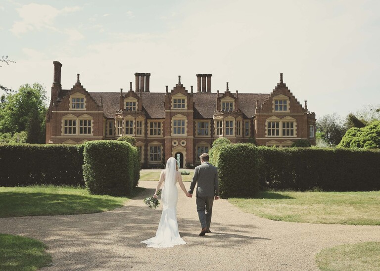 A bride and groom walking away holding hands with a magnificent hall in front of them on a wedding day at Haughley Park Barn photographed by suffolk wedding photographer Hayley Denston Photography