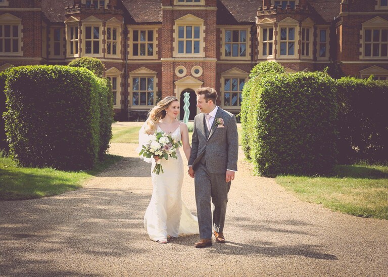 A bride and groom walking looking at each other holding hands with a red brick hall behind them on a wedding day at Haughley Park Barn photographed by suffolk wedding photographer Hayley Denston Photography
