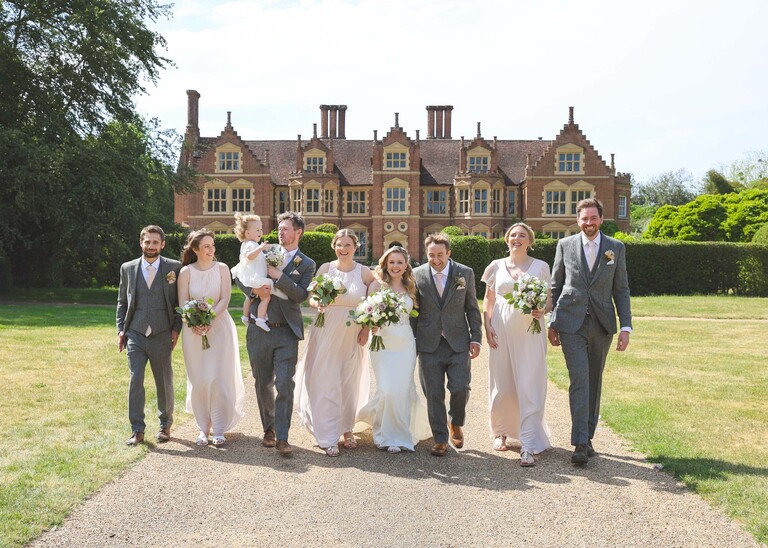 A bridal party in greys and pale pinks walking arm in arm for a group photo with a hall behind them on a wedding day at Haughley Park Barn photographed by suffolk wedding photographer Hayley Denston Photography