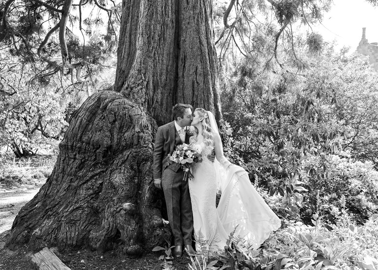 A black and white photo of a bride and groom stood in front of the base of a giant tree on a wedding day at Haughley Park Barn photographed by suffolk wedding photographer Hayley Denston Photography