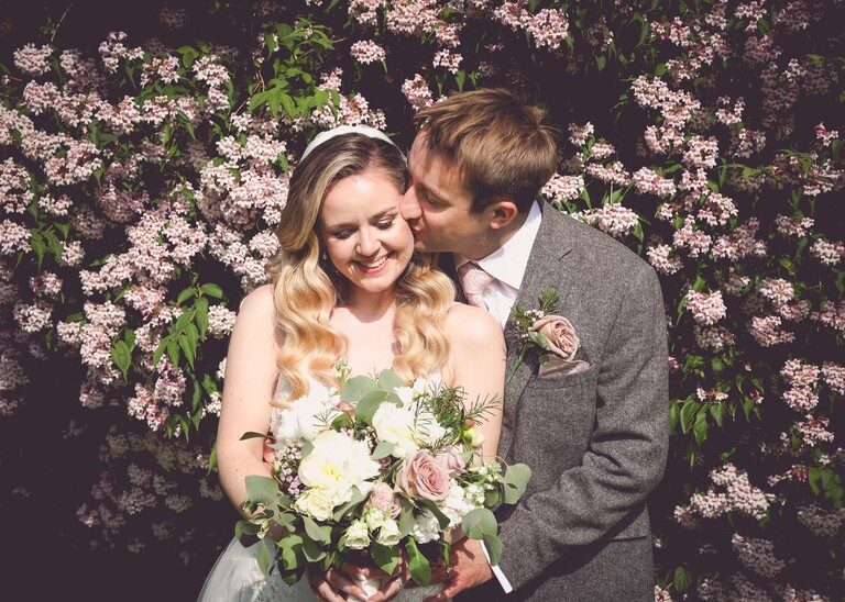 A groom kissing a bride on her cheek and cuddling her in front of a pale pink flowering bush on a wedding day at Haughley Park Barn photographed by suffolk wedding photographer Hayley Denston Photography