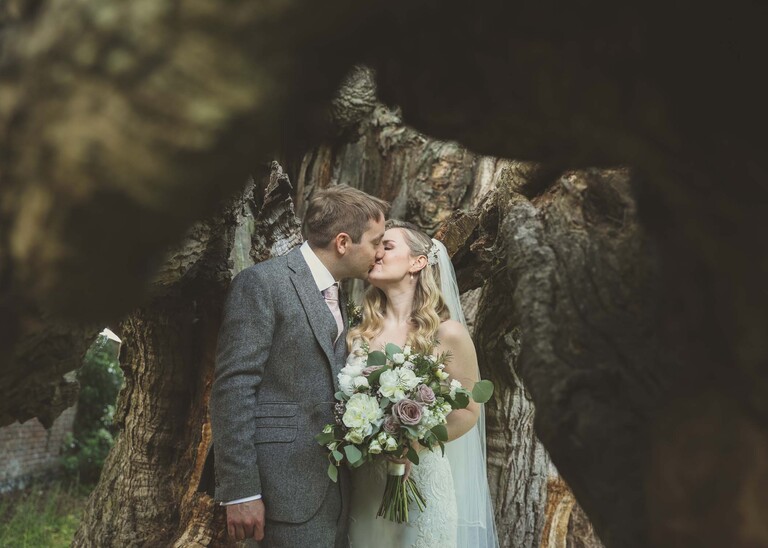 A bride and groom kissing through an old oak tree on a wedding day at Haughley Park Barn photographed by suffolk wedding photographer Hayley Denston Photography