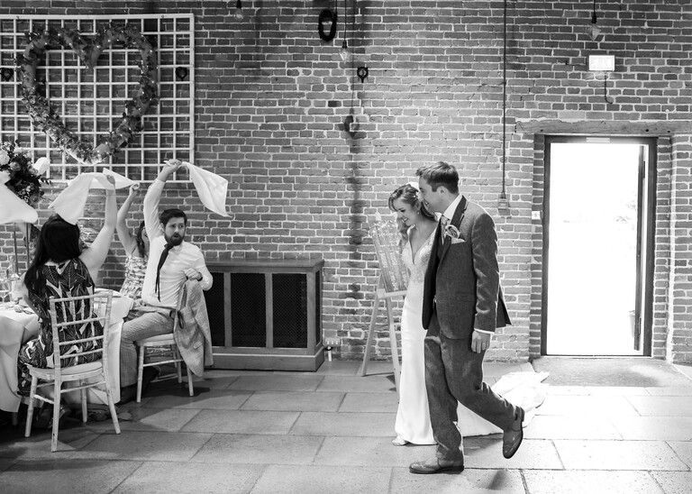 A black and white photo of a bride and groom walking in to their wedding breakfast with napkins being swung in the air by guests on a wedding day at Haughley Park Barn photographed by suffolk wedding photographer Hayley Denston Photography