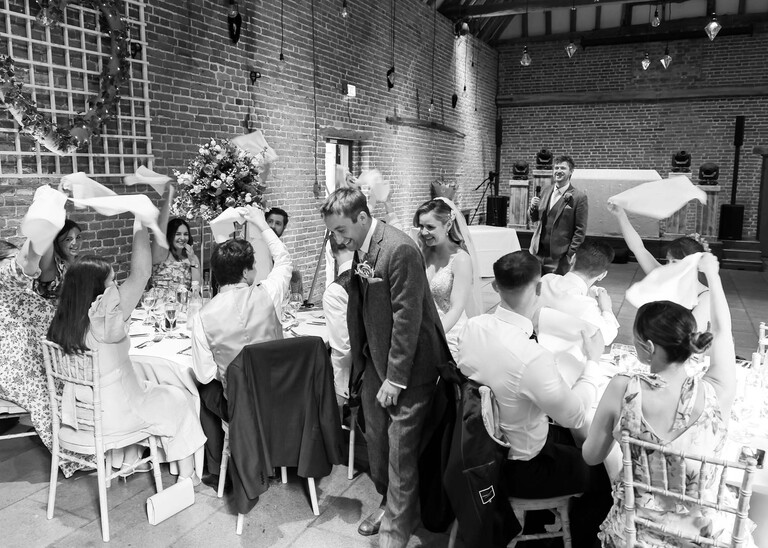 A black and white photo of a bride and groom walking in through tables for their wedding breakfast with guests waving napkins in the air on a wedding day at Haughley Park Barn photographed by suffolk wedding photographer Hayley Denston Photography