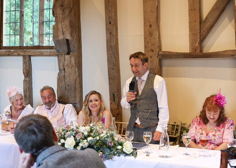 A groom giving his wedding speech with the bride and father of the bride laughing on a wedding day at Haughley Park Barn photographed by suffolk wedding photographer Hayley Denston Photography