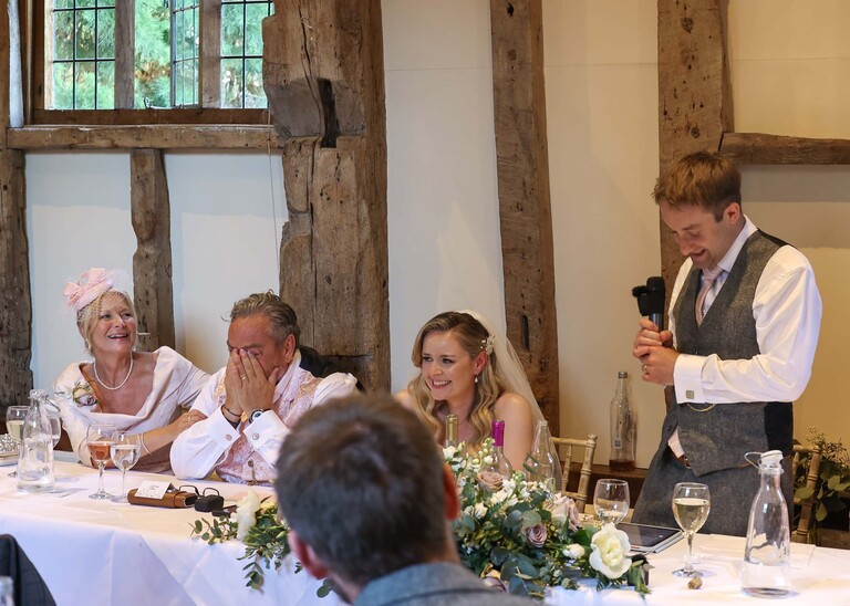 A groom giving his wedding speech with the bride laughing and the father of the bride crying on a wedding day at Haughley Park Barn photographed by suffolk wedding photographer Hayley Denston Photography