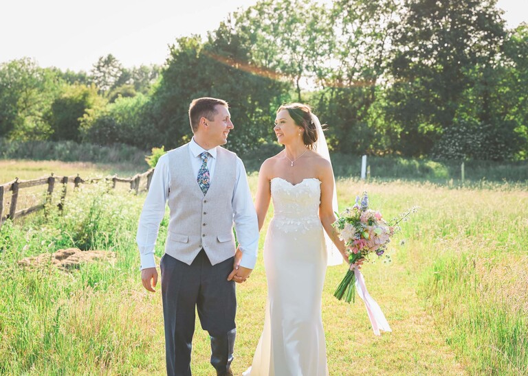 A bride and groom walking through a meadow holding hands looking at each other with the sun behind them on a wedding day at Little Green Wedding Barn photographed by suffolk wedding photographer Hayley Denston Photography