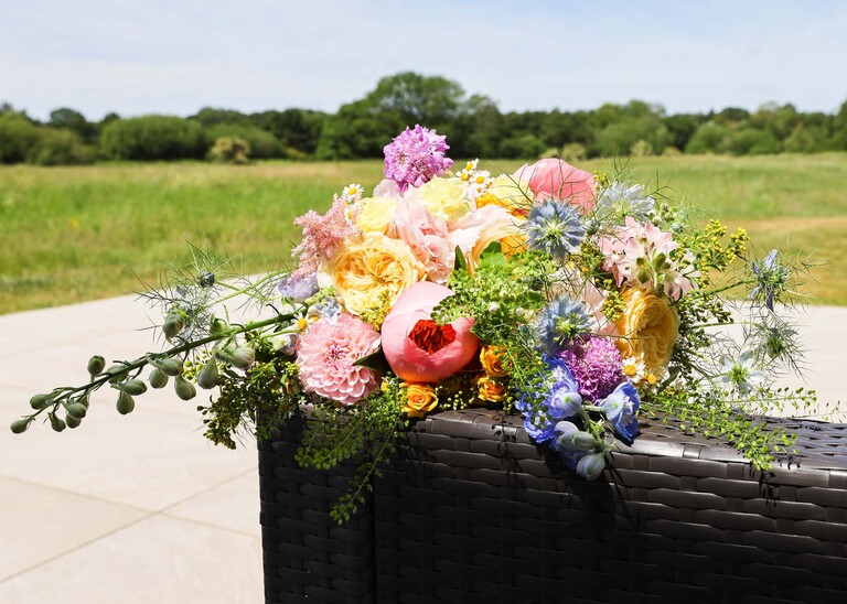 A bright floral bouquet sat out on a black rattan chair with beautiful meadows behind it on a wedding day at Little Green Wedding Barn photographed by suffolk wedding photographer Hayley Denston Photography