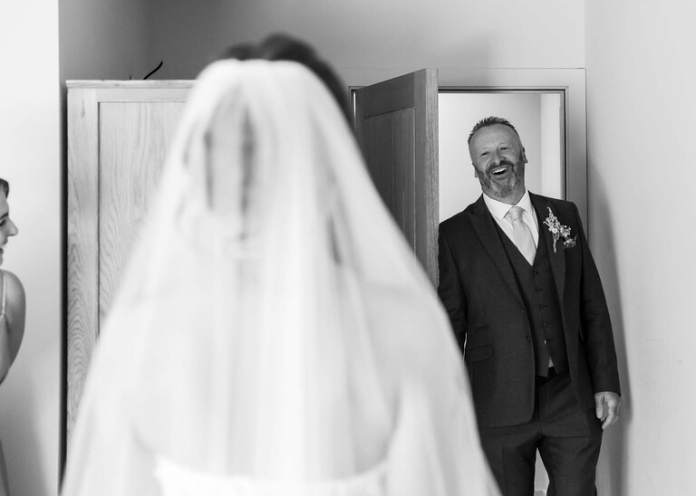 A black and white photo of a father of the bride coming in and seeing his daughter in her wedding dress for the first time on a wedding day at Little Green Wedding Barn photographed by suffolk wedding photographer Hayley Denston Photography