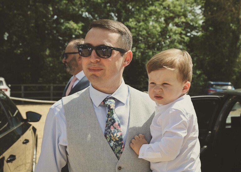 A groom with his shades on and holding his son arriving for his wedding day on a wedding day at Little Green Wedding Barn photographed by suffolk wedding photographer Hayley Denston Photography